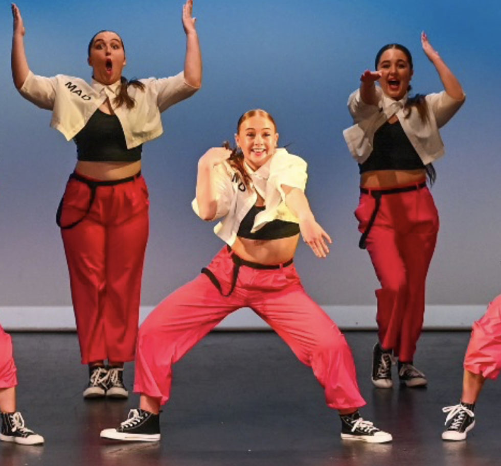 Hip Hop crew on stage. Three dancers are visible they wear pink pants with black trim a black top and a white short sleeved jacket. They are doing 3 different moves.
