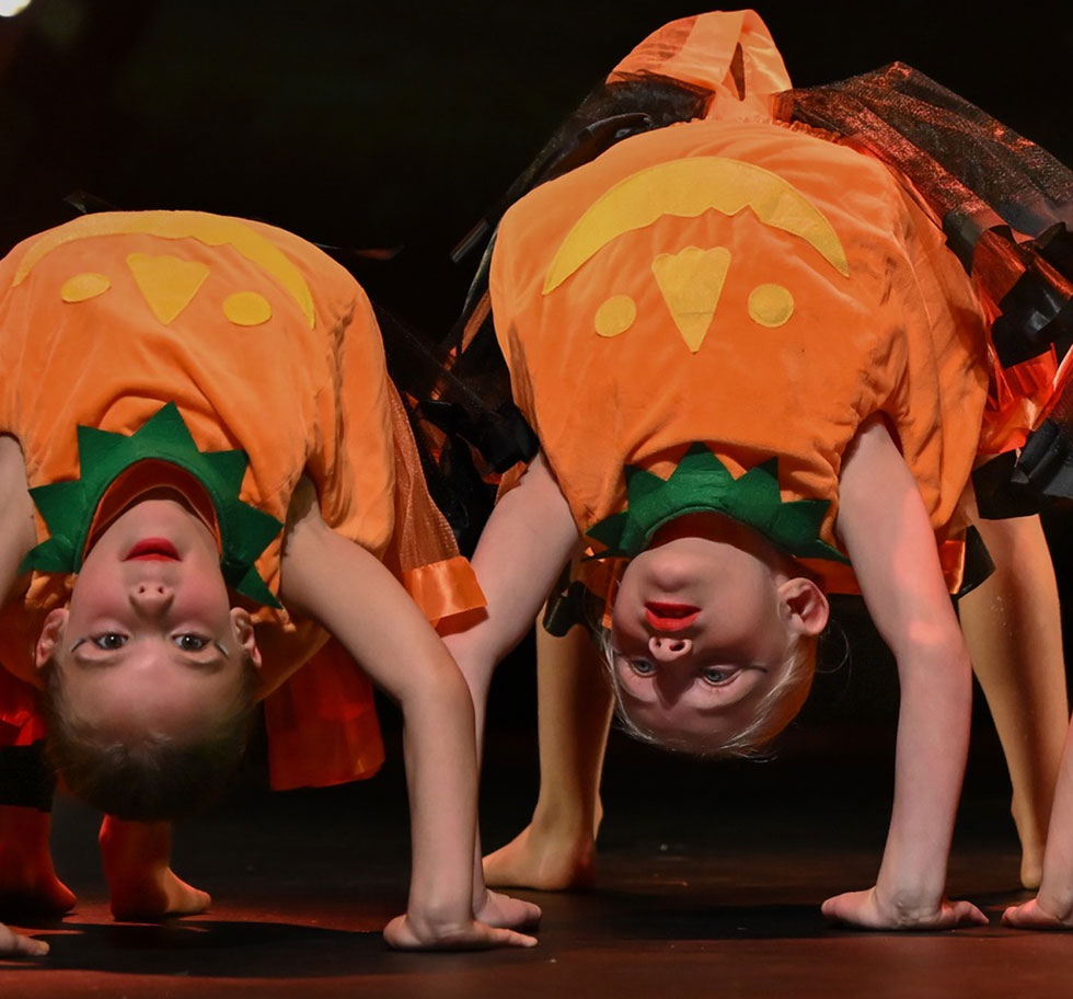 Two young ACRO dancers wearing pumpkin costumes arebent backwards with their hands and feet on the ground. They are looking at the camera.
