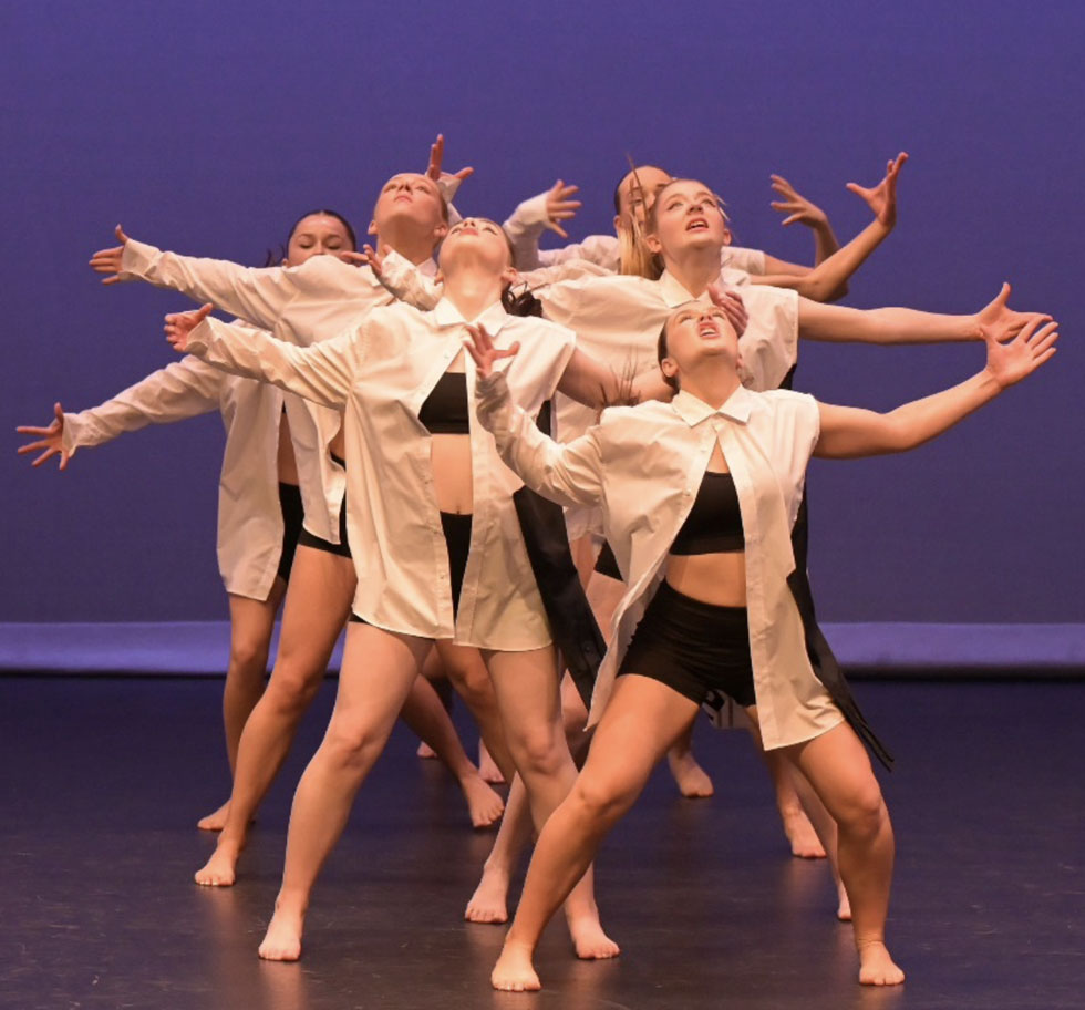 A contemporary dance group of 7 girls stand together with their arms spread and looking upwards. They wear beige oversized shirts over black tights
