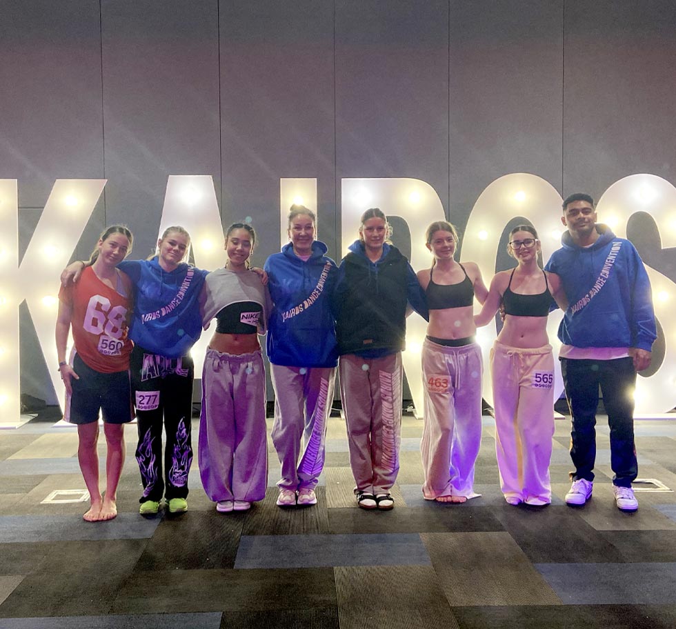 8 Dancers stand in front of a large lighted sign the reads KAIROS
