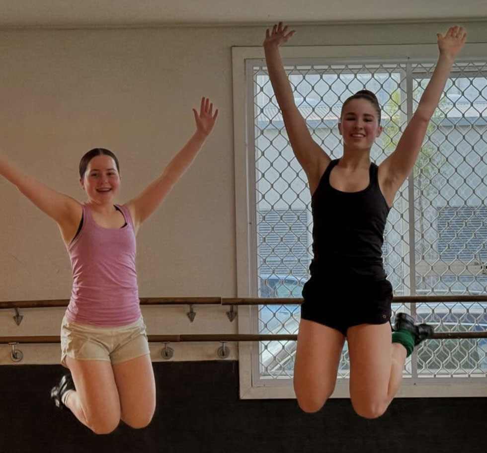 Two dancers jump in the air with arms up and feet tucked up high behind them. They are in a dnace studio and wear casual active wear.