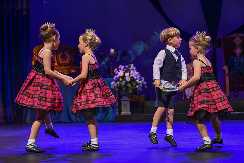 Four young dancers perform highland dance. Three girls wear a red tartan quilt. The Boy wears shorts and a vest.