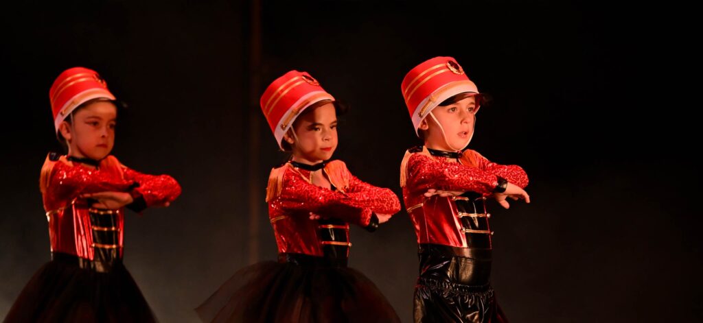 Three young dancers permorming on stage. The wear soldier costumes with red jackets and matching red hats with black pants/skirts.