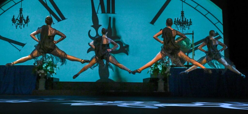 Four dancers look away from the audience while performing jumping splits. There is a large blue clock face behind them.